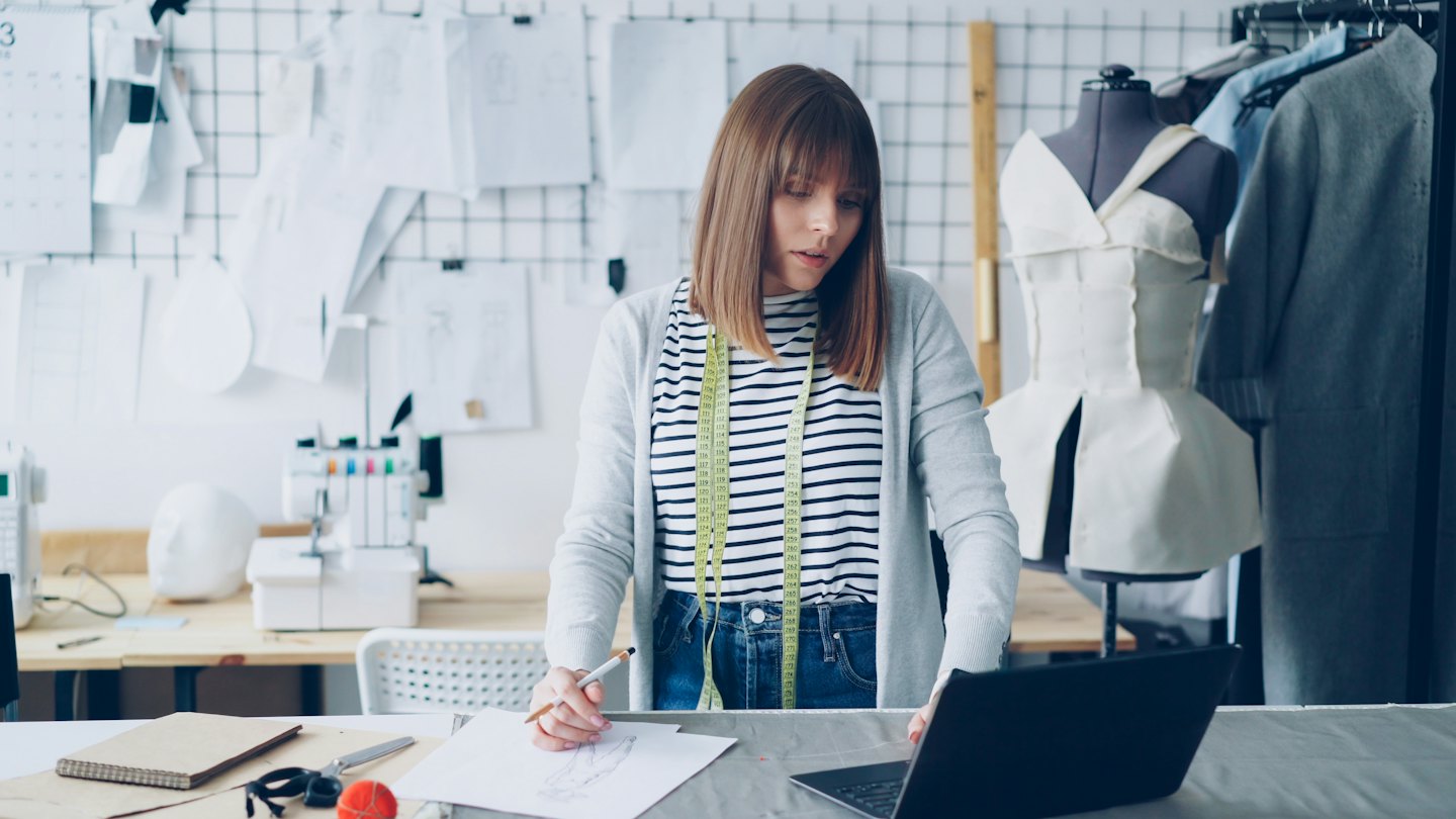 Fashion designer sketches at a desk with a laptop and patterns.