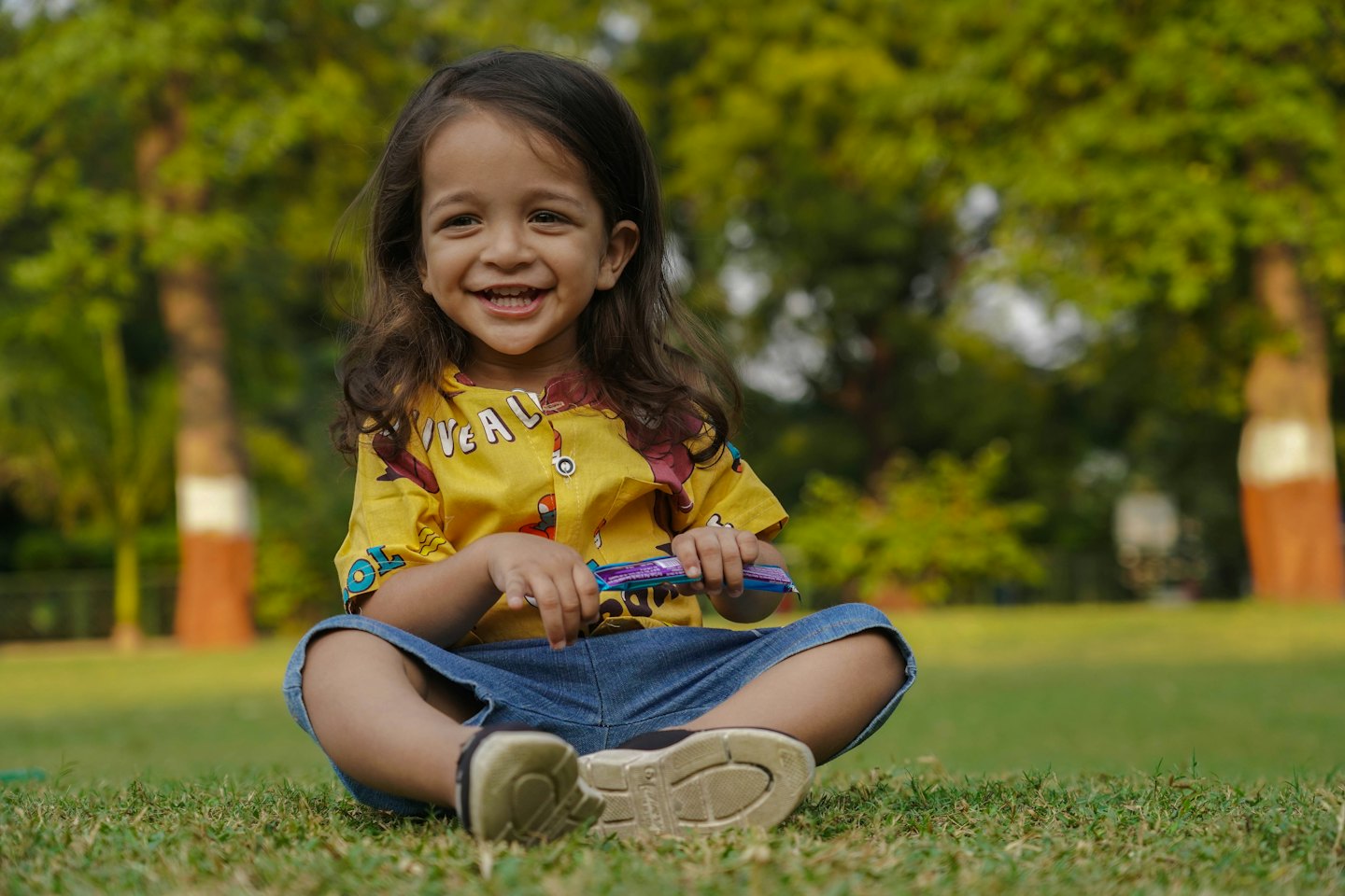 Child in yellow shirt sits cross-legged on grass holding a purple packet.